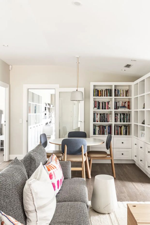 Bright, compact open-plan living and dining area with a grey sectional sofa in the foreground, a round dining table with upholstered timber chairs, and extensive white built-in bookshelves and cabinetry along the rear wall. Pale walls and ceiling, dark timber flooring, a simple pendant light, and abundant books create a clean, inviting residential interior.