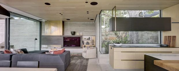 Open-plan contemporary living and kitchen interior with floor-to-ceiling glazing, a pale timber ceiling, minimalist cabinetry, a large island, and a dark sectional sofa. The living area includes a built-in fireplace or media feature, while the kitchen has light wood cabinetry and a contrasting dark suspended rangehood volume. Natural vegetation is visible outside the glazing.