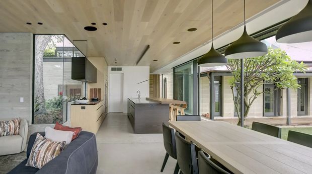 Open-plan contemporary kitchen and dining area with a large timber dining table, integrated kitchen cabinetry and island, pendant lighting, extensive floor-to-ceiling glazing, and a leafy courtyard visible beyond. A sofa occupies the foreground, creating a connected living zone. The room features pale timber ceiling boards, light flooring, dark-framed glazing, and a calm indoor-outdoor connection.