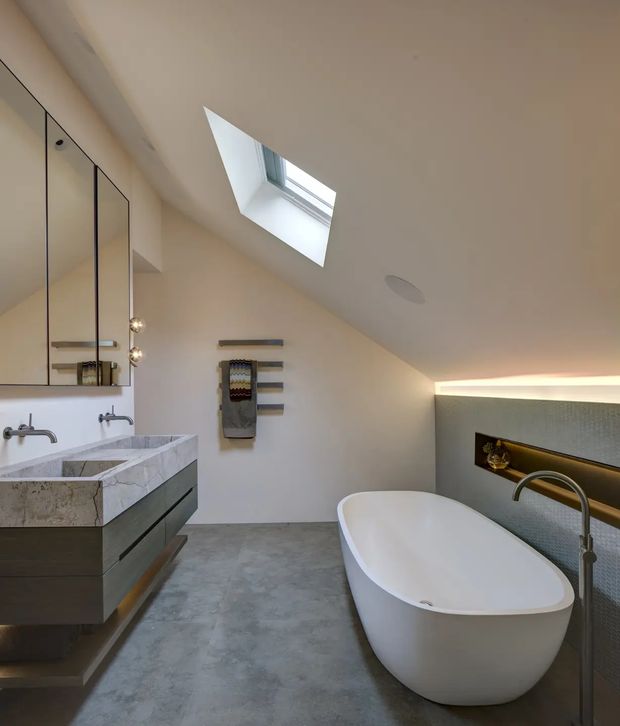Contemporary bathroom with a freestanding white bathtub, double vanity, wall-mounted mirrors, skylight, and a recessed feature niche with integrated lighting. The room has a sloping ceiling, grey concrete-look flooring, a stone vanity benchtop, and muted neutral finishes.