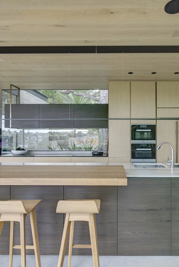 A contemporary kitchen with extensive light timber cabinetry, a broad timber island with integrated seating, dark wood-grain lower cabinetry, stainless-steel appliances, and a white undermount sink with a curved chrome tap. Floor-to-ceiling glazing and dark roller blinds overlook greenery and neighbouring homes. The ceiling features timber lining and a dark linear recessed detail with recessed downlights.