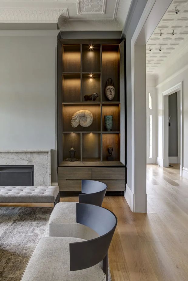 A refined living room with a tall, dark timber built-in display cabinet illuminated by integrated spotlights and filled with sculptural objects. The room features ornate decorative ceiling detailing, pale walls and trim, a marble fireplace surround, warm timber flooring, a textured neutral rug, and contemporary curved-back lounge chairs and ottomans. An adjoining bright hallway is visible through a wide opening.