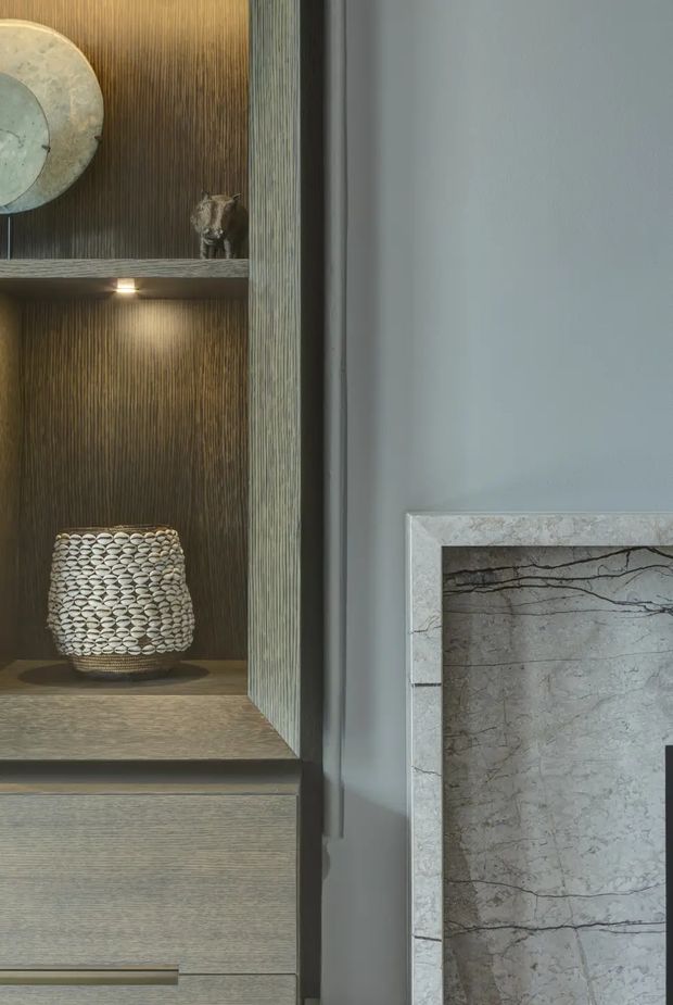 Close-up interior detail showing a built-in timber display cabinet with vertically fluted dark wood backing, integrated shelf lighting, decorative objects, and lower drawers. To the right is a pale grey wall with a marble-clad fireplace surround and insert. The composition is refined, muted, and material-focused.