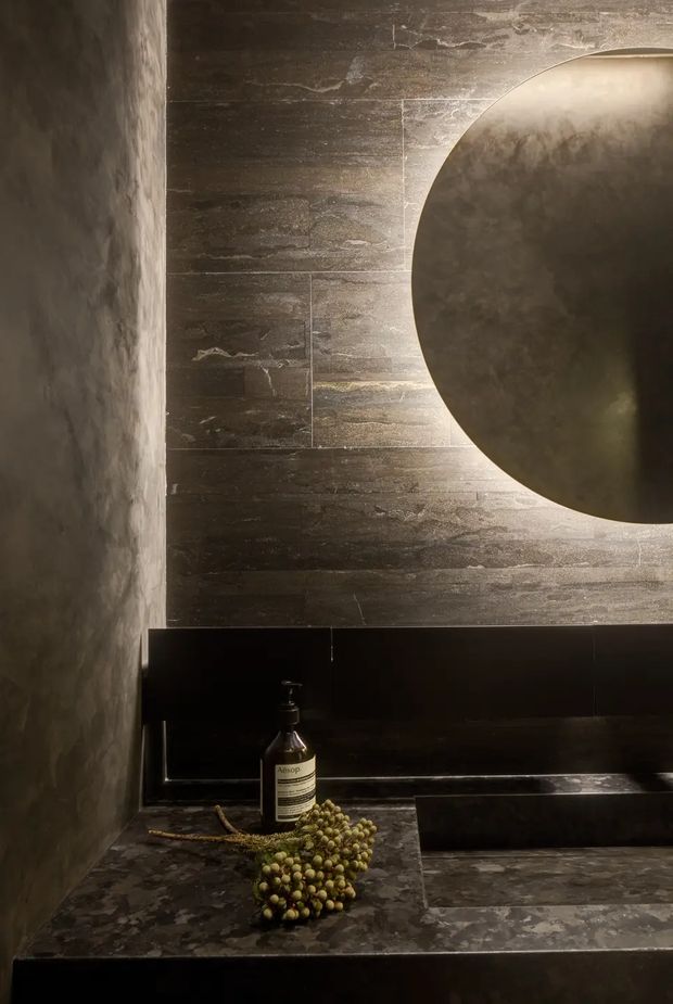 A dark, atmospheric bathroom vanity vignette with a large round wall mirror, textured dark stone or plaster wall surfaces, black built-in basin cabinetry, and a mottled stone benchtop. Warm concealed lighting outlines the mirror and highlights the tactile finishes. An Aesop soap dispenser and decorative greenery sit on the vanity.