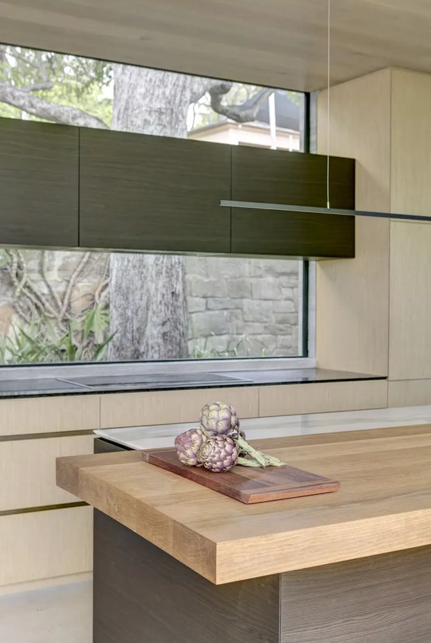 A refined contemporary kitchen focused on a large timber island with a thick light-oak benchtop and dark wood-grain cabinetry. Pale wood wall cabinetry and a long horizontal window bring natural light into the space, while a slim linear pendant is suspended above the island. A dark stone or composite worktop runs beneath the window, with a garden and masonry wall visible outside.