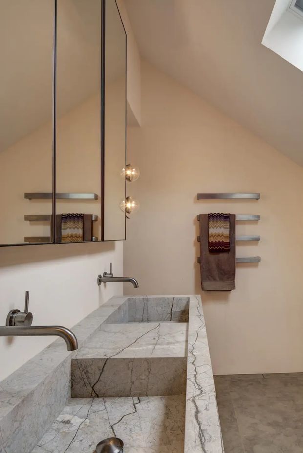 A refined contemporary bathroom with a double vanity clad in prominently veined pale natural stone. Two wall-mounted metal faucets and basins sit beneath a large mirrored cabinet, while sculptural spherical pendant lights hang in front of the mirror. Minimal horizontal towel rails with folded towels are mounted on the warm off-white walls. The room has a sloped ceiling with a skylight and a grey stone or concrete-look floor.