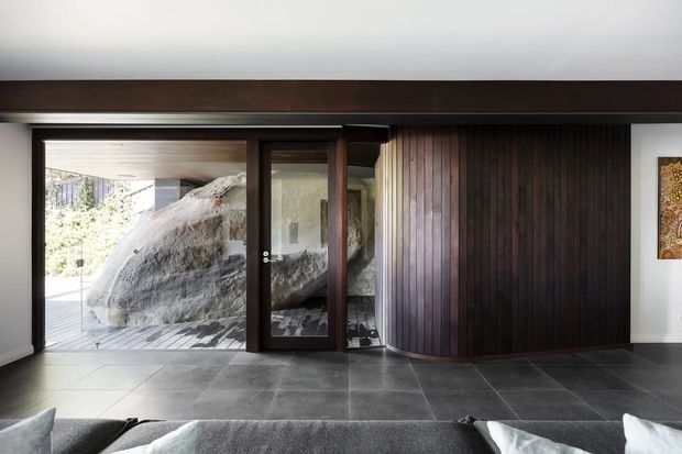 Interior view of a contemporary living room or entry area with dark large-format tiled flooring, a dramatic curved wall clad in vertical dark timber battens, and extensive glazing with a dark timber-framed door. A large natural rock formation is visible immediately outside beneath a covered terrace. The room has a restrained, high-contrast palette and strong indoor-outdoor connection.