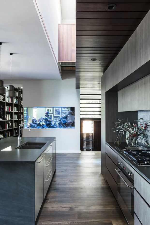 A contemporary galley-style kitchen with a large dark island, integrated double sink and minimalist tapware. Dark timber flooring continues through the space, while full-height cabinetry and a built-in cooktop and oven line the right wall. A timber-lined ceiling with recessed downlights leads toward a bright glazed opening at the rear. Open shelving, pendant lights, and a wall-mounted aquarium or artwork add visual interest.