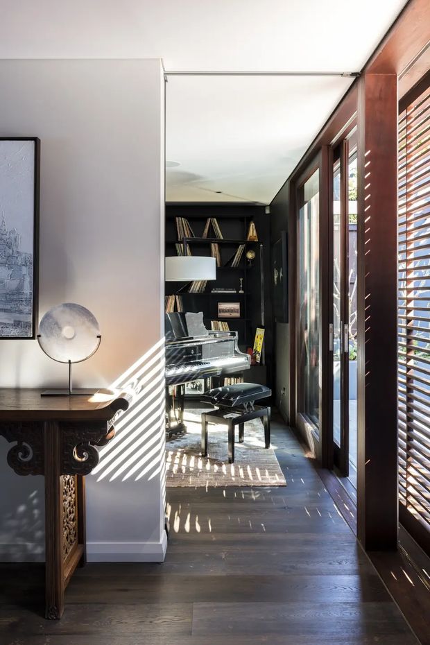 A narrow, sunlit interior passage leading to a compact music room. The space features dark timber flooring, black built-in shelving filled with records or books, a piano with bench, tall timber-framed glazing and timber louvred shutters on the right, and a richly carved dark wood console table on the left. Strong striped sunlight and shadows create a dramatic, refined atmosphere.