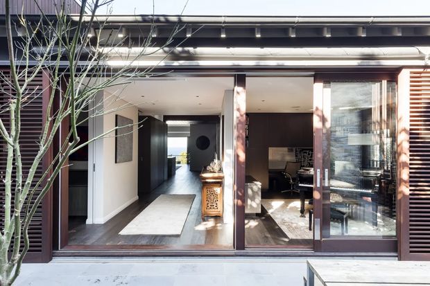A modern residential facade viewed from outside, with wide glazed sliding doors opening onto a dark timber-floored interior. The entrance leads through a hallway toward a bright rear opening, while a compact home office or study is visible behind the glazing on the right. The exterior features dark timber framing, horizontal timber screening, a projecting roof edge, and a pale paved forecourt.