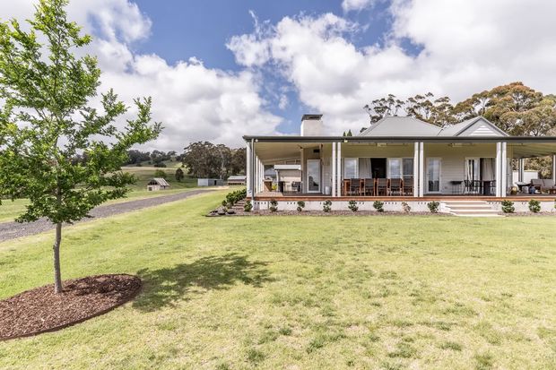 Exterior view of a low-rise rural home set within expansive landscaped lawns and open countryside. The house has a light rendered facade, pale pitched roofs with gabled elements, broad covered verandas, slender posts, extensive glazing, and outdoor dining areas. A gravel driveway and mature trees reinforce the spacious rural setting.