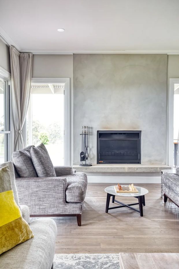 Bright, refined living room with a large textured grey plaster fireplace wall, recessed gas fireplace and pale stone hearth. The room has light timber flooring, neutral upholstered armchairs and sofas, floor-length curtains, tall windows, and a small round coffee table. A yellow accent cushion and muted patterned rug add subtle colour.