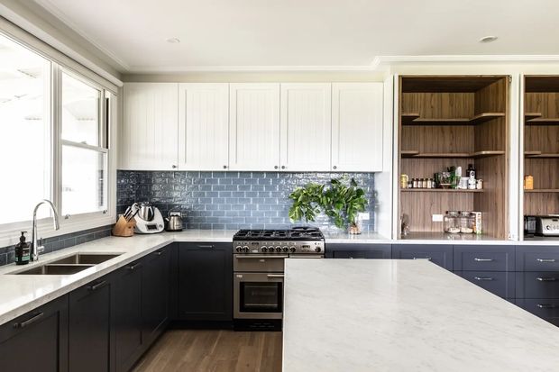 Bright contemporary kitchen with a large white stone island, dark lower cabinetry, white vertical-panel upper cabinets, a blue-grey glossy brick-tile splashback, timber open shelving, stainless steel appliances, and timber flooring. Natural light enters through a large window above the sink.