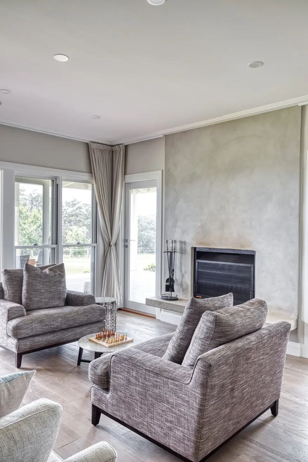 A bright, refined living room with two upholstered armchairs and a sofa arranged around a small coffee table with a chess set. The room features a built-in dark fireplace set into a textured pale grey wall, full-height glazed doors and windows opening to an outdoor area, layered curtains, recessed ceiling lights, white crown moulding, and light timber flooring.