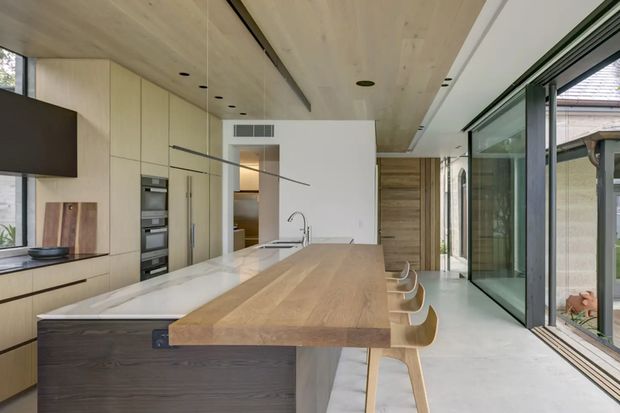 A bright, open-plan contemporary kitchen with a long timber-topped island and integrated breakfast bar seating. Pale timber cabinetry runs along the left wall with built-in ovens and concealed storage, while a white stone work surface and undermount sink occupy the island. Full-height dark-framed glazing opens the kitchen toward an exterior space, and a pale timber-lined ceiling with recessed lighting adds warmth.