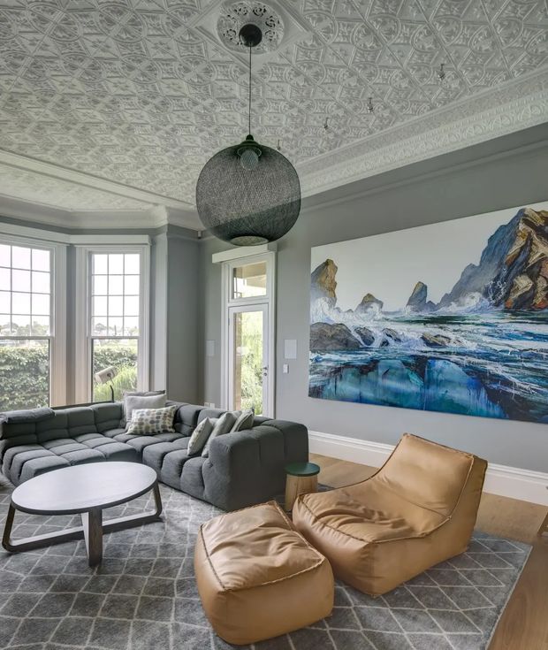 A refined living room with grey walls, tall bay windows, an ornate pressed-pattern ceiling and decorative cornice, a large woven pendant light, a deep grey sectional sofa, round coffee table, tan leather lounge chair and ottoman, and a large blue-toned mountain and seascape artwork. The room combines traditional architectural detailing with contemporary furnishings.