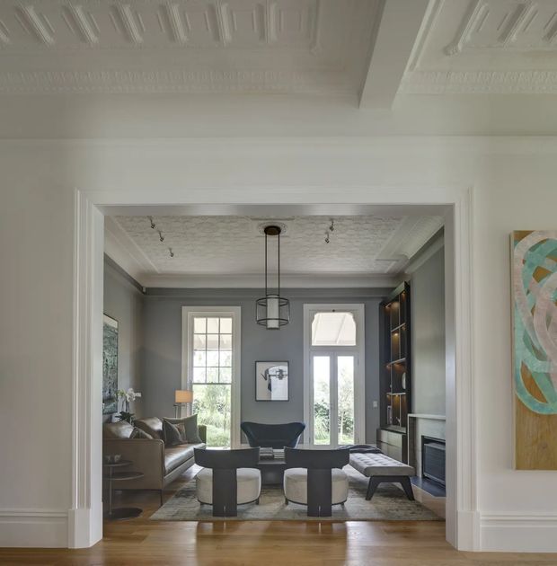 A refined living room viewed through a wide trimmed opening, featuring grey walls, dark built-in shelving and fireplace joinery, a brown sofa, sculptural lounge chairs, ottomans, and a central coffee table. Tall glazed doors and windows bring in garden light. The room has polished timber flooring, elaborate white decorative ceiling detailing, substantial wall mouldings, and a contemporary pendant light, blending heritage architectural elements with sophisticated modern furnishings.