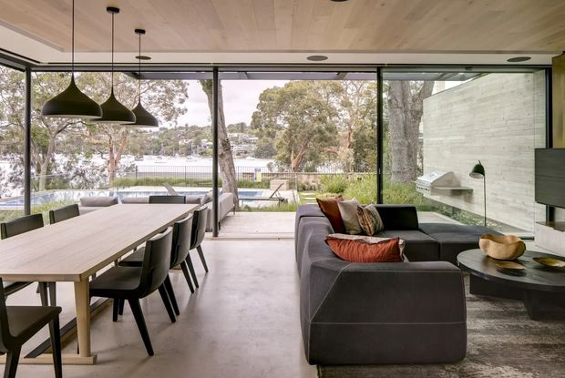 Open-plan contemporary living and dining area with full-height glazing overlooking a leafy suburban landscape and pool area. A long timber dining table with dark upholstered chairs sits beneath sculptural pendant lights, while a large charcoal sectional sofa faces a wall-mounted television and minimalist fireplace. The room features a pale timber-panelled ceiling, polished concrete flooring, dark window frames, and a pale concrete or stone feature wall.