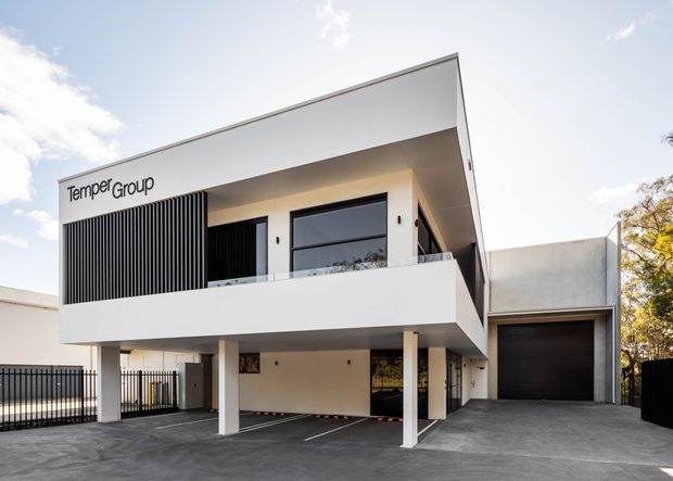 A modern multi-storey commercial or mixed-use building exterior with a crisp white geometric facade, broad projecting upper-level balcony, black vertical screening, large glazed openings, and an attached enclosed garage or service bay. The ground level is supported by slender columns and opens onto a paved parking or driveway area. The facade bears the text “Tempe Group.”