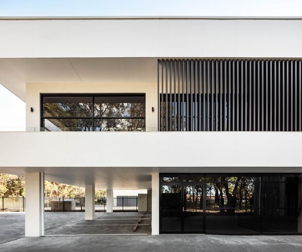 A contemporary multi-level building exterior with a crisp white rendered facade, deep covered ground-level area, dark-framed glazing, and a prominent upper-level screen of closely spaced vertical black fins. The lower level includes an open sheltered parking or entry zone, while trees and fencing are visible through the glazing and around the building.