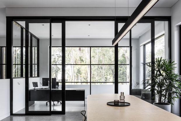 A bright contemporary open-plan workspace and meeting area with a large light-toned table in the foreground, black-framed glass partitions enclosing a compact office, expansive windows overlooking trees, and a linear suspended pendant light. Indoor planting and restrained black-and-white finishes add a polished, professional feel.
