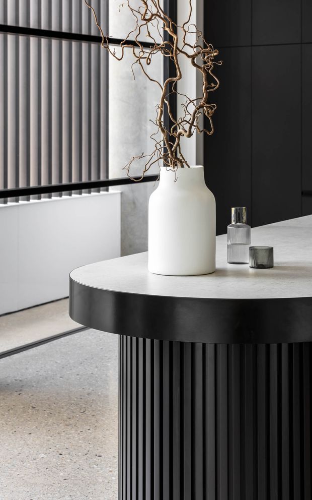 A close-up interior view of a refined contemporary space featuring a round, dark fluted built-in counter or island with a pale stone benchtop. A sculptural white ceramic vase with dried branches and small glass vessels sit on the surface. Vertical black ribbed wall panelling, a dark framed window with vertical blinds, a pale concrete-like wall, and a terrazzo-style floor create a monochromatic, textural setting.