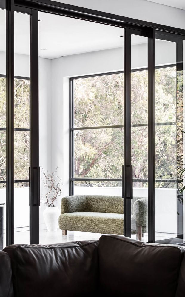 A bright, minimalist interior featuring full-height black-framed glazed doors or partitions, white walls and ceiling, and large windows overlooking leafy trees. A compact olive-grey upholstered sofa sits in the adjoining light-filled space, while the foreground contains a dark leather sofa. The strong black framing and expansive glazing create a crisp, contemporary architectural composition.