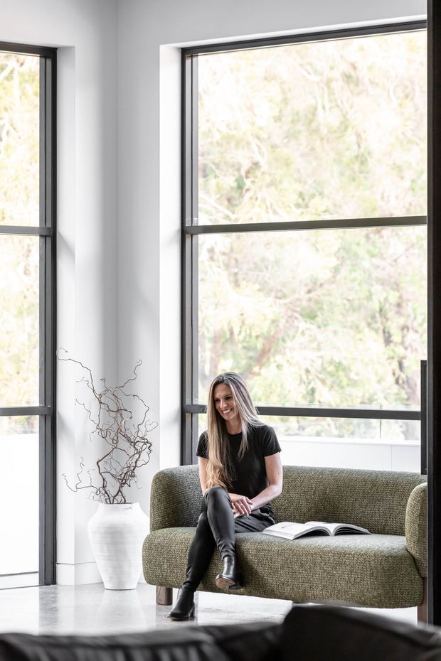 Bright contemporary living area with full-height black-framed windows, white walls, and polished pale flooring. An olive-green textured sofa sits beside a large white ceramic vessel with sculptural branches, while a person reads on the sofa. The space has a clean, refined, nature-connected feel with abundant daylight.