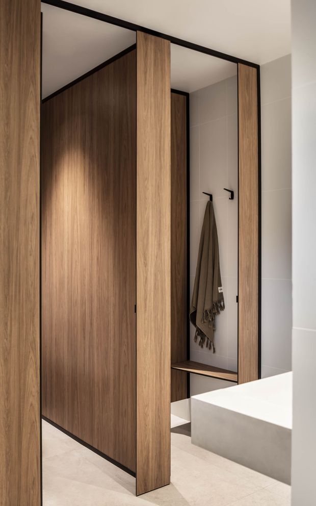 Contemporary bathroom with a warm timber-lined open wardrobe or entry niche, black-framed panels, pale large-format wall and floor tiles, wall hooks, and a large white rectangular freestanding or built-in bathtub visible in the foreground. The design is clean, refined, and minimalist with integrated timber joinery and concealed storage.