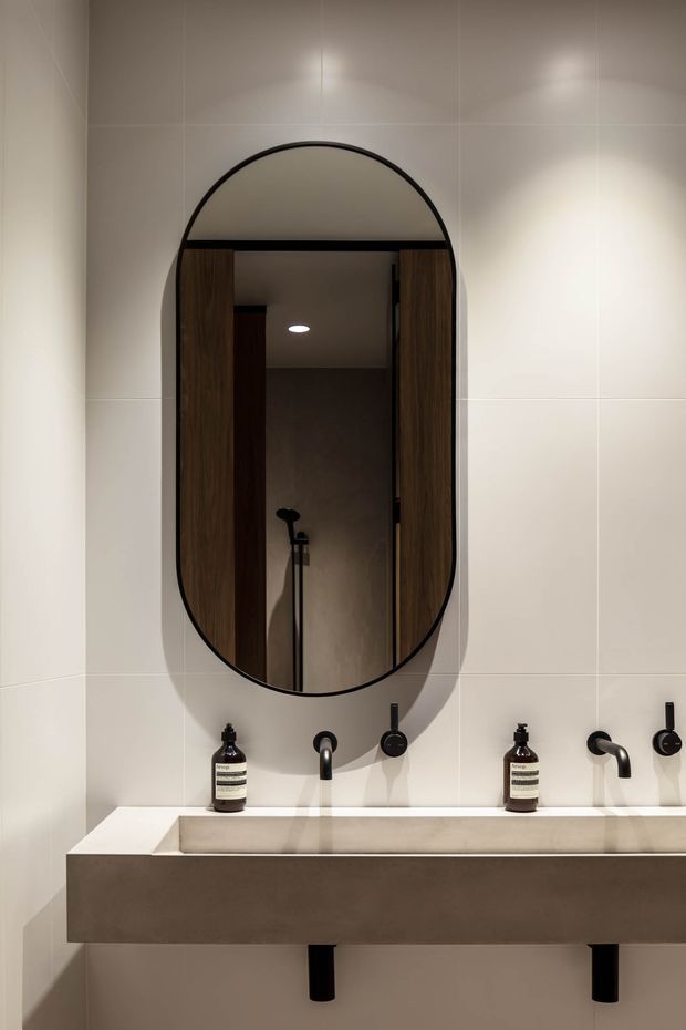 Contemporary bathroom vanity with a long wall-mounted rectangular basin, twin black wall-mounted faucets, dark timber cabinetry accents, and an elongated arched mirror with a black frame. Large pale tiled walls and soft integrated lighting create a refined, warm neutral interior.
