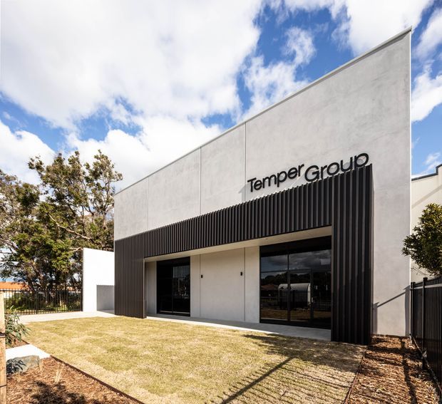 Street-facing view of a contemporary commercial building with a stark geometric facade, pale concrete or rendered wall panels, and a deep black vertical-slat entrance surround. Large glazed doors and windows open onto a small landscaped lawn, with the Temper Group sign mounted above the entrance. Bright daylight and partly cloudy sky are visible.