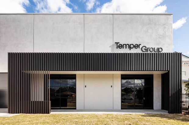 Street-facing commercial building facade with a minimalist rectangular form, smooth light-grey upper panels, a black vertical fin screen framing the entrance, dark glazed doors and windows, and the TemperGroup logo mounted above. The low-rise building is photographed from outside under a partly cloudy sky.