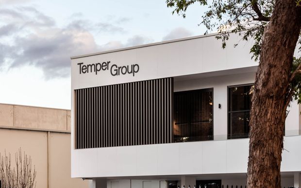 Exterior view of a contemporary commercial building for TemperGroup. The white, rectilinear facade features a projecting upper volume, vertical dark screening fins, large glazed openings, and integrated business signage, partially framed by a mature tree.