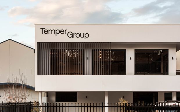 Front exterior view of a contemporary multi-storey commercial or office building with a clean white rectangular facade, dark recessed glazing, vertical metal screening, and a prominent TemperGroup sign. A black metal fence and adjacent building are visible in the foreground and background.