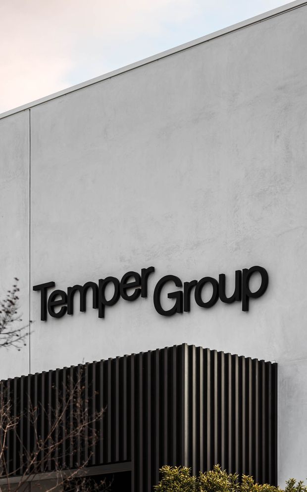 Exterior architectural photograph of a contemporary commercial building facade. A large smooth light-grey rendered or concrete wall carries raised black “Temper Group” signage, with a black vertical slatted screen forming a projecting lower facade element and sparse landscaping at the base.