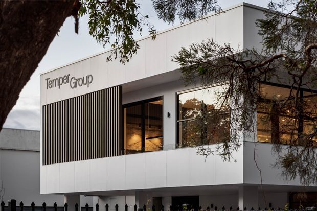 Contemporary two-storey commercial building with a crisp white rectangular facade, extensive dark-framed glazing, and a vertical timber-look screening element. The building is photographed externally through mature trees, with illuminated interior spaces visible behind the windows and signage reading “Tamper Group.”