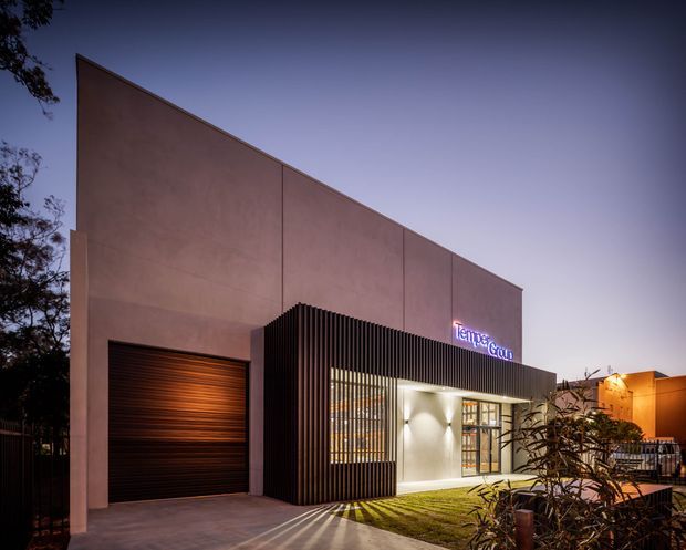 Exterior dusk photograph of a contemporary commercial building with a tall angular parapet, smooth muted terracotta facade, dark ribbed screening, a roller garage-style door, illuminated entrance glazing, and a glowing business sign. Landscaping and a paved approach sit in front of the building.