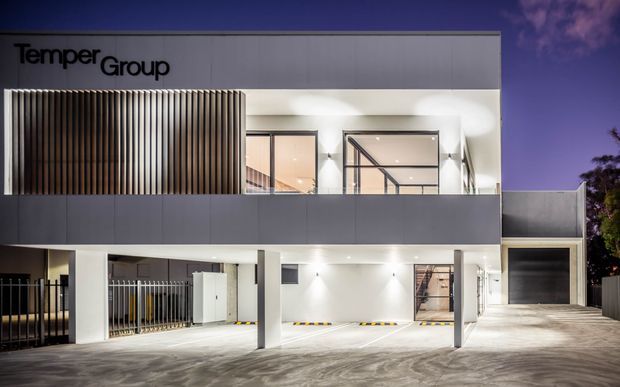 Night-time exterior photograph of a contemporary multi-level commercial building branded “TemperGroup”. The facade combines smooth pale-grey panels, extensive glazing, vertical timber-look screening, exposed concrete columns, open ground-level parking, and a recessed garage or loading bay. Bright architectural lighting highlights the clean geometric form and entrance areas.