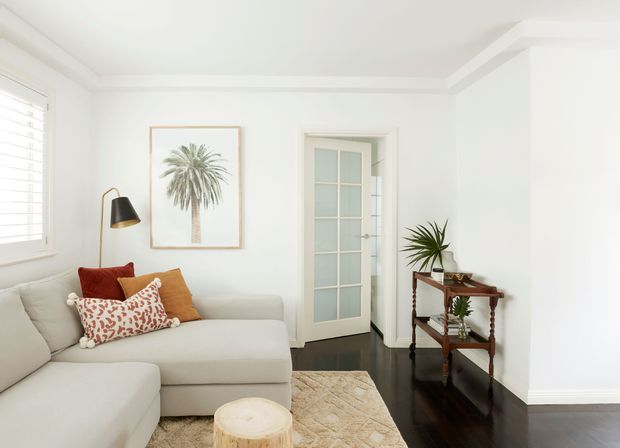 Bright, airy living room with a large cream sectional sofa, patterned and rust-toned cushions, a black-and-brass floor lamp, palm artwork, a small timber console table with plants, a textured rug, and dark polished timber flooring. White walls feature decorative ceiling coving and trim, with a glazed French-style door leading to another room.