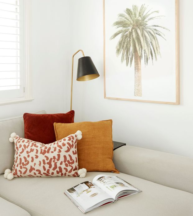 Bright, softly styled living room corner featuring a pale upholstered sofa, layered rust, ochre, and patterned cushions, a slim brass-and-black floor lamp, a large framed palm artwork, and a light timber-framed window with white shutters. An open design magazine rests on the sofa, creating a relaxed, curated atmosphere.