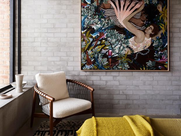 A moody, art-filled interior corner featuring a mid-century-style timber and woven lounge chair beside a dark ledge, set against a white-painted brick wall. A large colourful botanical artwork provides a strong focal point, while a mustard-yellow throw and patterned rug add warmth and contrast.
