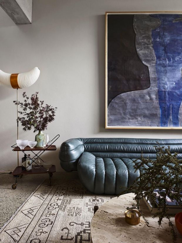 A moody, artfully styled living room featuring a deep blue-grey sculptural leather sofa, a round pale stone coffee table, a patterned vintage-style rug, a timber-and-metal drinks trolley, a sculptural floor lamp, greenery, and a large abstract artwork in a slim timber frame. The space has a refined mid-century-inspired character with strong organic forms and dark, muted tones.