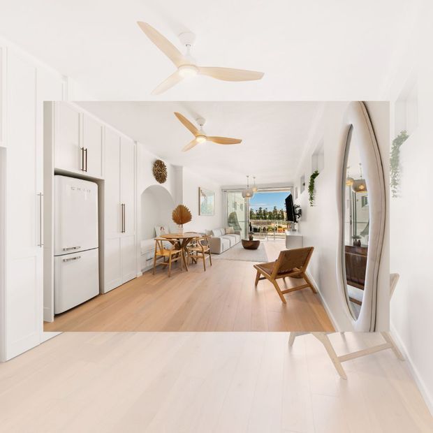 Bright open-plan interior combining a white galley kitchen with integrated cabinetry and appliances, a compact dining area, and a living room opening to an outdoor terrace. Pale timber flooring, white walls and joinery, sculptural arched niches, woven and timber furniture, ceiling fans, greenery, and a large arched mirror create an airy coastal-inspired scheme.