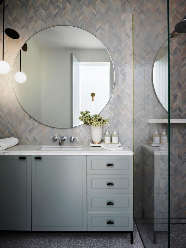 A refined contemporary bathroom featuring a grey vanity with integrated basin, black hardware, a large round mirror, patterned grey chevron wall tiles, terrazzo-like flooring, and a frameless glass shower enclosure. Soft globe lighting and restrained styling create a sophisticated, calm atmosphere.