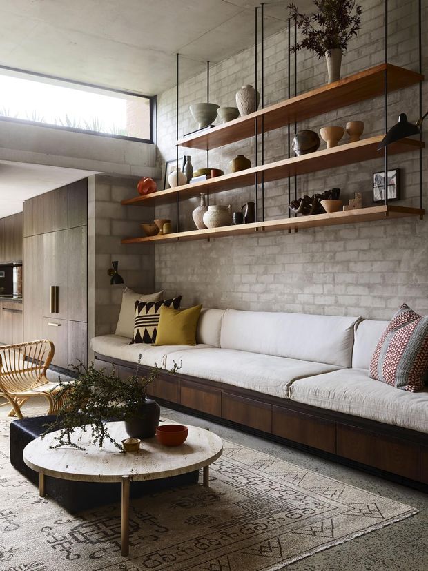A warm, contemporary living room with a long built-in upholstered sofa, dark timber storage base, floating timber shelves on black metal supports, and a textured exposed concrete-block feature wall. Neutral furnishings are accented by mustard and patterned cushions, while a round light stone coffee table, woven rug, pottery, and indoor planting add organic character. A compact kitchen or joinery area is visible at the left.