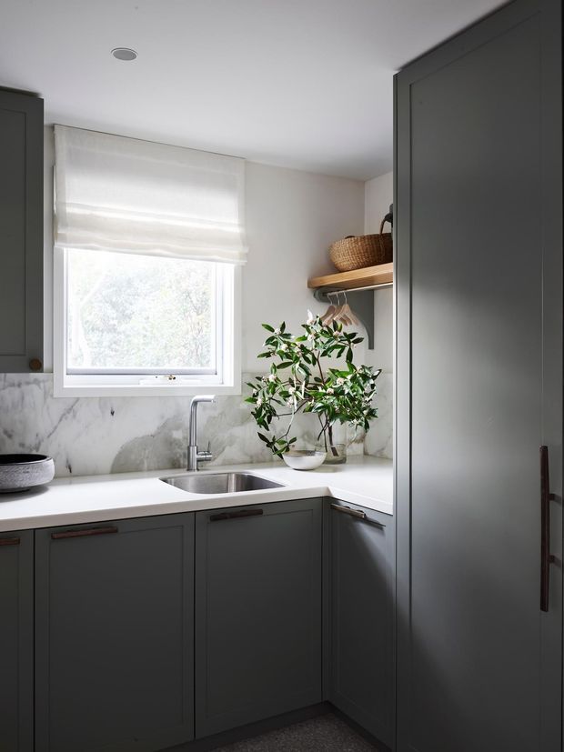 Compact kitchen corner with dark grey shaker-style cabinetry, white benchtops, a stainless-steel undermount sink and chrome tap, marble-look wall finish, and a bright window with a white Roman blind. Open timber shelving with a woven basket and hanging rail adds warmth, while a potted plant sits beside the sink.