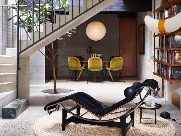Open-plan mid-century-inspired interior with a concrete stairway and black metal balustrade crossing above a dining area. The space features exposed grey brick walls, polished light terrazzo flooring, timber built-in bookshelves, a dark timber door, mustard upholstered dining chairs, a sculptural black chaise lounge, and a large spherical pendant light. Indoor planting and layered furniture create a warm, eclectic living and dining environment.