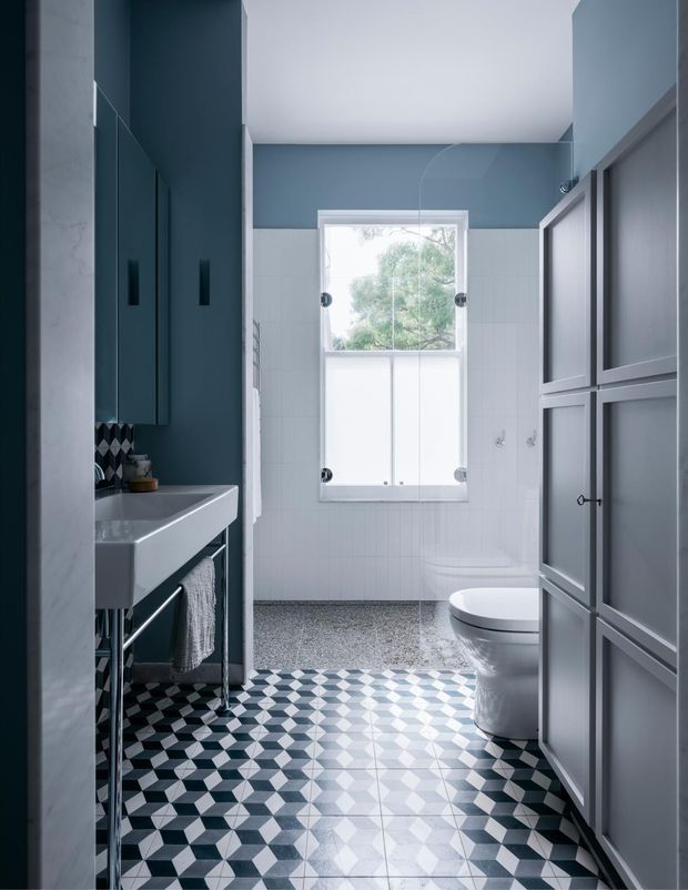 A stylish contemporary bathroom viewed from the entrance, featuring a white pedestal-style basin with dark metal legs, dark blue-grey wall cabinetry, patterned black, white and grey geometric floor tiles, white wall tiles, a frosted-glass shower enclosure, and a wall-hung toilet. A tall window provides natural light, while dark framed built-in cabinetry lines the right side.