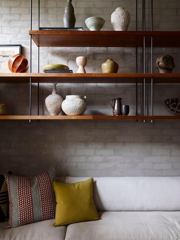 A moody, styled living room vignette featuring a white upholstered sofa beneath three long timber shelves with black metal supports. The shelves display sculptural ceramic and wooden vessels, books, and decorative objects against a textured exposed brick wall. Patterned and mustard cushions add warmth and contrast.