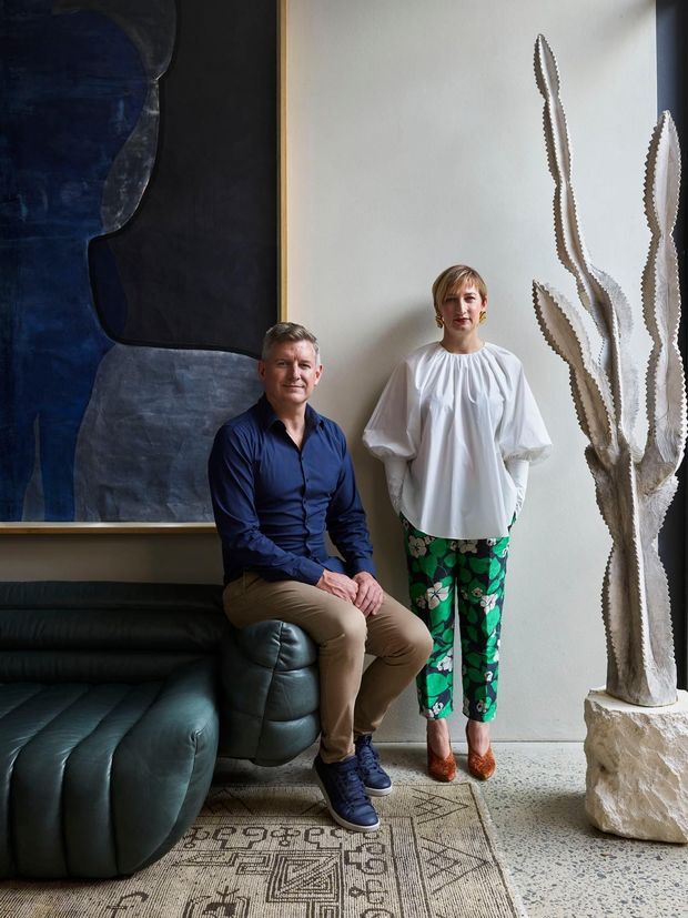 Portrait of two people posed in a refined contemporary interior, beside a large dark abstract artwork, a deep green leather sectional sofa, and a tall sculptural cactus-like artwork on a stone plinth. The room features pale walls, terrazzo flooring, and a patterned area rug.